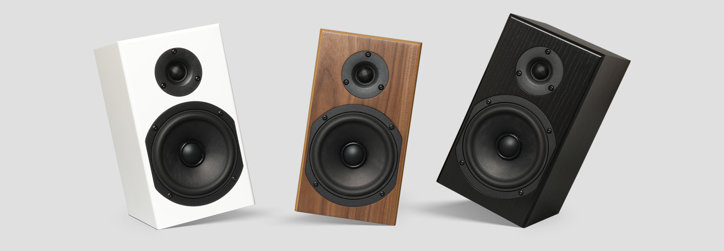 The three Loon finishes side by side with grilles off - satin white, black walnut, black ash - each tilted back to show the big black woofer and the small dome tweeter above it