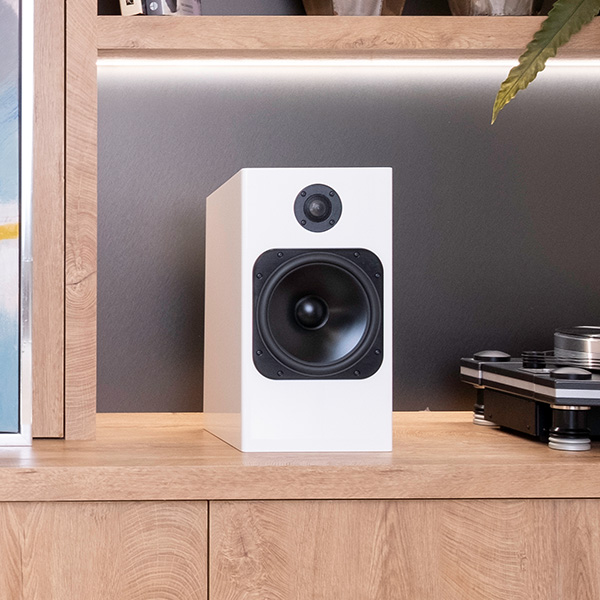 Element Fire V2 in Ice gloss white on a light oak sideboard beside a turntable and an amplifier, a lit shelf behind it and a plant leaf overhead