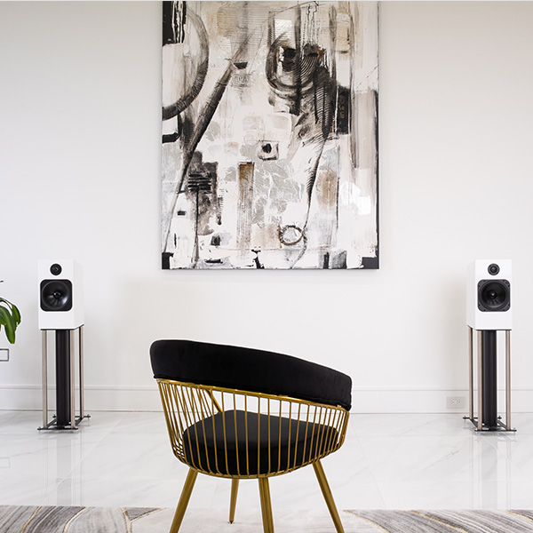 A pair of Element Fire V2 in Ice gloss white on stands either side of a large abstract painting, with a gold-framed velvet chair on a patterned rug in front